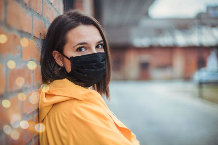 Chica de pelo corto apoyada contra una pared con una sudadera amarilla y una máscara negra