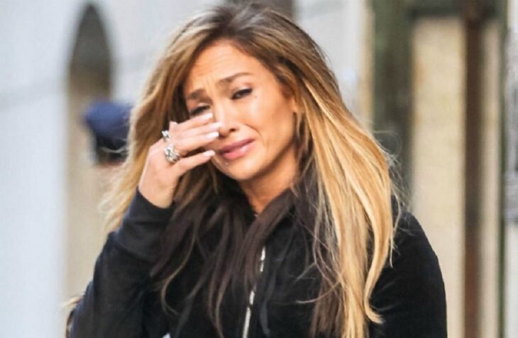 Imagen de la cantante Jennifer Lopez llorando, tiene el cabello rubio suelto y esponjoso y viste una sudadera negra, tiene anillos brillantes en la mano derecha y se seca las lagrimas 