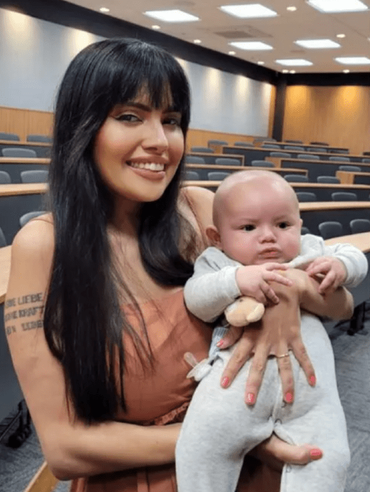 una madre joven sostiene a su bebé en brazos dentro de un aula de la universidad; ella lleva un vestido color arena sin mangas y el bebé un mameluco gris