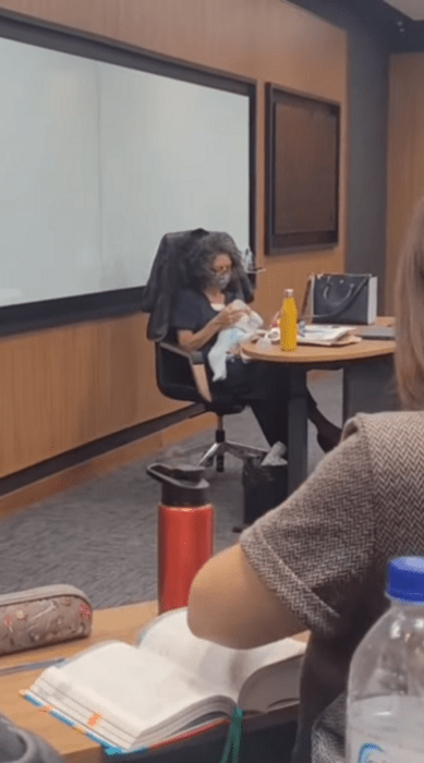 una profesora alimenta con un biberón a un bebé mientras está sentada frente a su escritorio, durante un examen que realiza a sus alumnos, lleva ropa de color oscuro