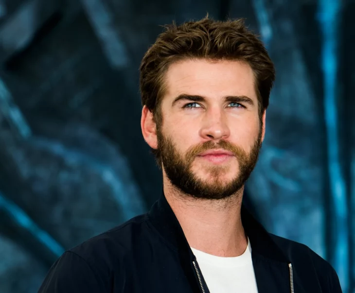 Liam Hemsworth en una sesión de fotos, lleva un saco negro con una camiseta blanca, resaltando el azul de sus ojos frente a un fondo oscuro, con barba y bigote