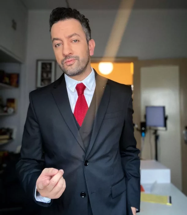 Imagen del comediante y youtuber Chumel Torres, lleva un traje negro con corbata roja y camisa blanca, tiene el cabello corto y una sombra de barba y bigote.