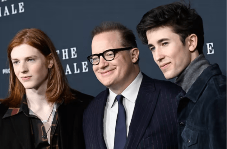 Brendan Fraser posando junto a sus hijos, viste un traje azul marino con camisa blanca y corbata azul, mientras que los chicos llevan vestimenta casual, uno tiene el cabello pelirrojo y el otro negro.