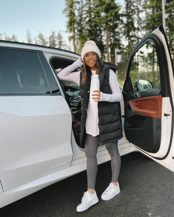 foto de una mujer posando recargada en una camioneta rodeada de árboles usando un chaleco holgado y un gorro blanco