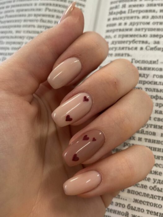 Uñas color nude con una uña en color café y diseños de corazón