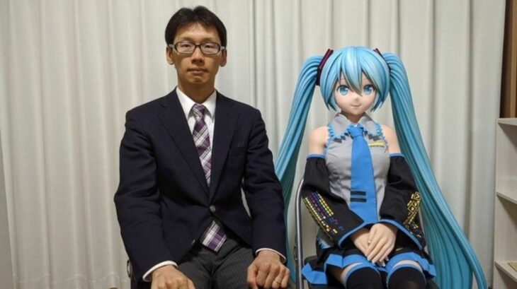 Akihiko Kondo sentado al lado de una muñeca de Miku, un personaje ficticio 