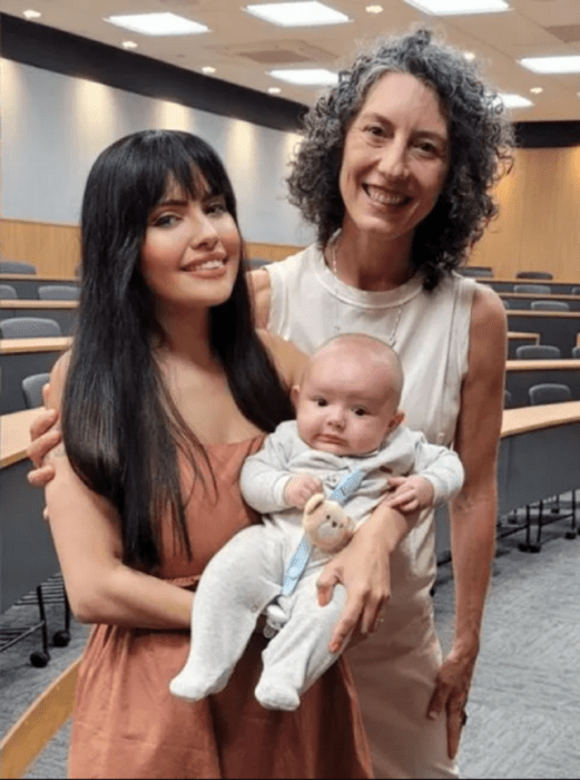 imagen dentro del aula de una Universidad, dos mujeres posan en la foto, una sostiene a un bebé en brazos; las tres llevan ropa casual