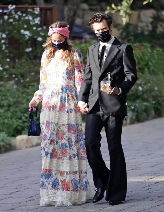Harry Styles y Olivia Wilde caminando tomados de la mano, ella lleva un vestido casual largo con estampado floreado y una banda rosa sobre la cabeza, él porta un traje negro con camisa blanca y corbata de moño negra, ambos usan cubrebocas negros