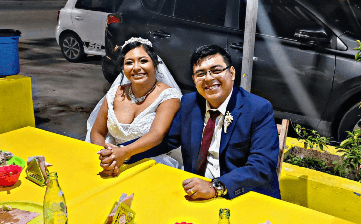 dos recién casados sentados en una mesa de un puesto de tacos, tomados de la mano y sonriendo felices