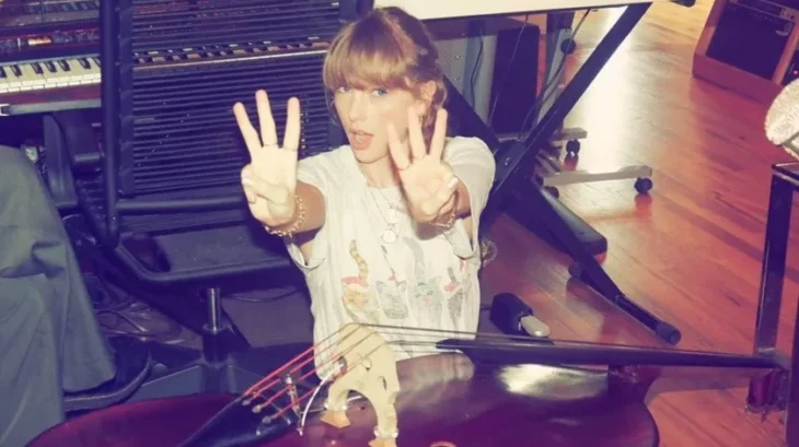 Taylor Swift mostrando un tres con sus dedos en cada mano, al lado de un piano