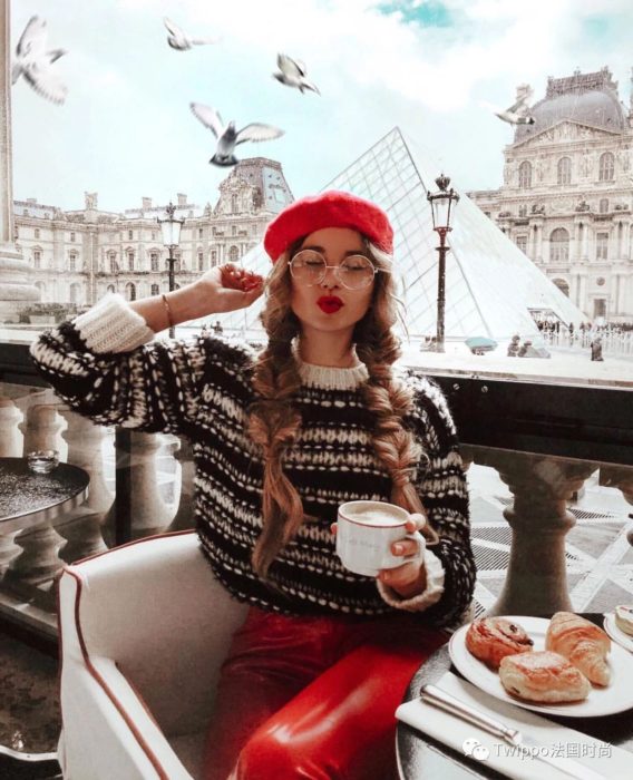 Chica sentada en un café de París usando un feo suéter navideño mientras disfruta de un café