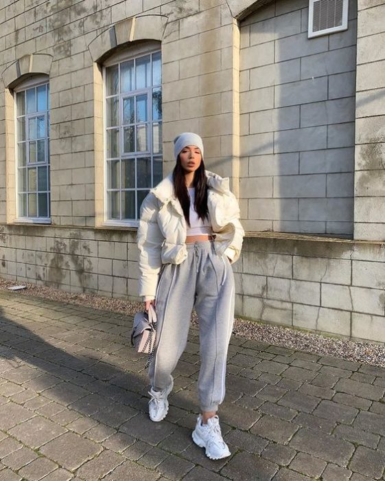 chica de cabello castaño largo usando una gorra gris, camisa blanca, abrigo blanco, pantalones deportivos grises, tenis blancos y bolsa de mano gris