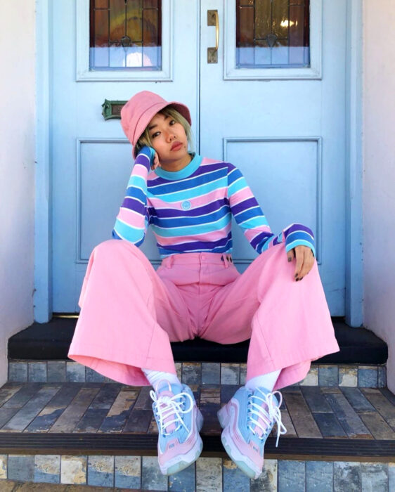 Look colorido aesthetic; mujer coreana de cabello corto y rubio sentada en escaleras frente a una puerta azul, vestida con colores vibrantes, gorro pescador rosa, suéter rayado en morado, azul fuerte y claro, pantalón acampanado y tenis chunky en tonos pastel
