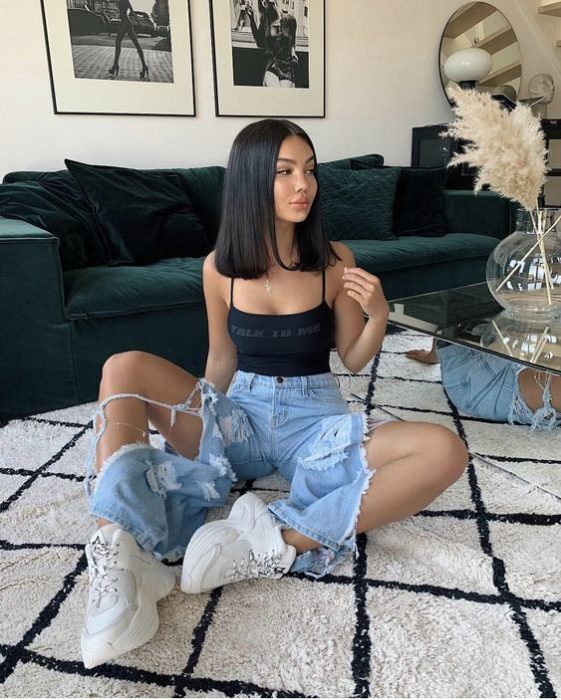 Chica morena de cabello negro lacio sentada en el piso con jeans rotos y blusa de tirantes negra