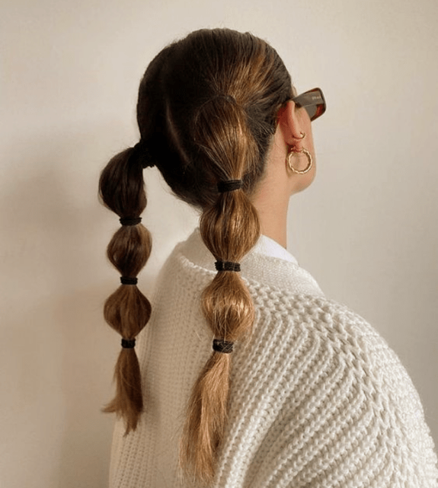 mujer de cabello claro y largo con coletas abombadas, usando un suéter blanco y gafas de sol negras