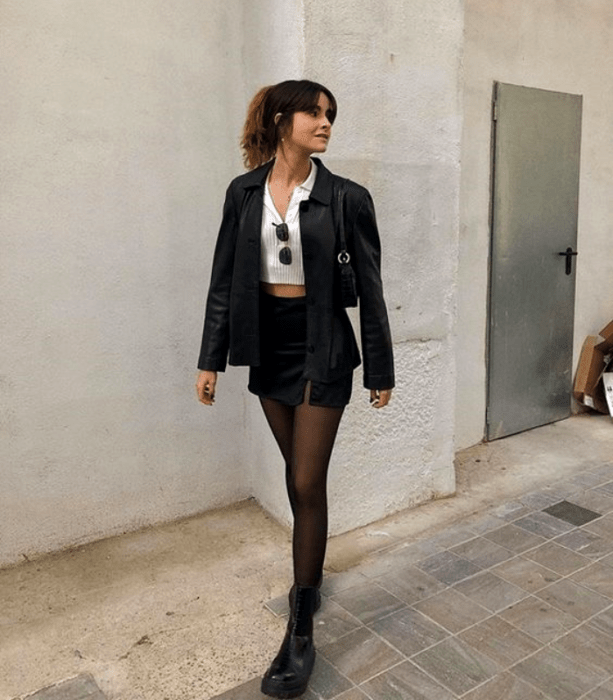 chica con cabello castaño usando un top blanco, gafas de sol, chaqueta de cuero, minifalda negra y medias negras
