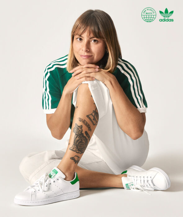 Línea de ropa y calzado de Adidas, Stan Smith Disney; sudadera color hueso con estampado verde de la Rana René; mujer sentada en el suelo, sonriendo, con cabello rubio castaño y un tatuaje de gato en la pierna