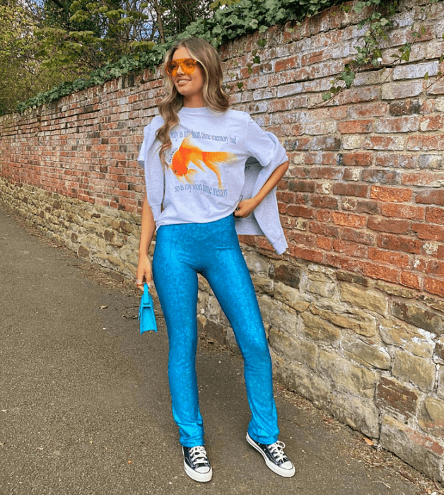 chica de cabello claro con lentes de sol naranja, camiseta gráfica blanca, pantalones azul brillante, tenis converse negros, bolso pequeño celeste