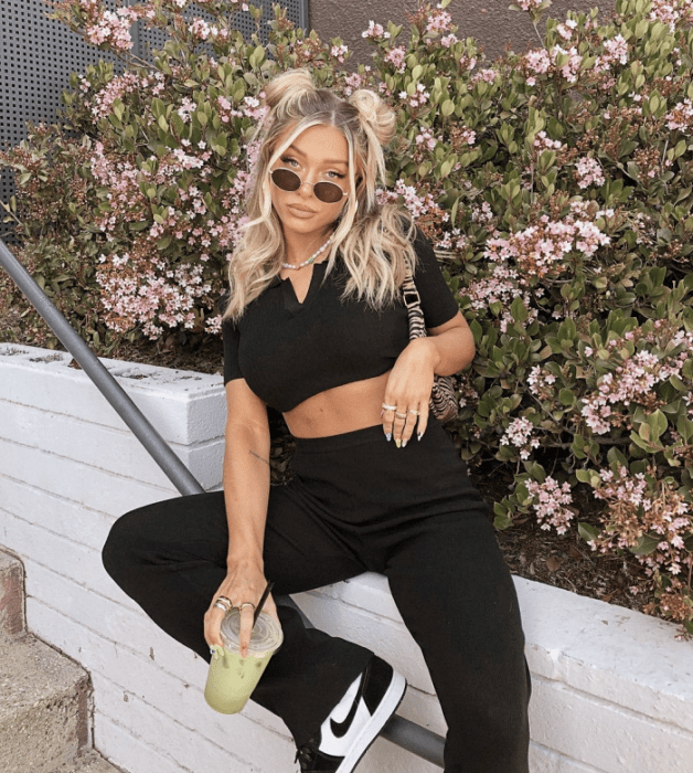 chica de cabello rubio usando lentes de sol, crop top negro de manga corta, pantalones negros ajustados acampanados, tenis deportivos blancos con negro