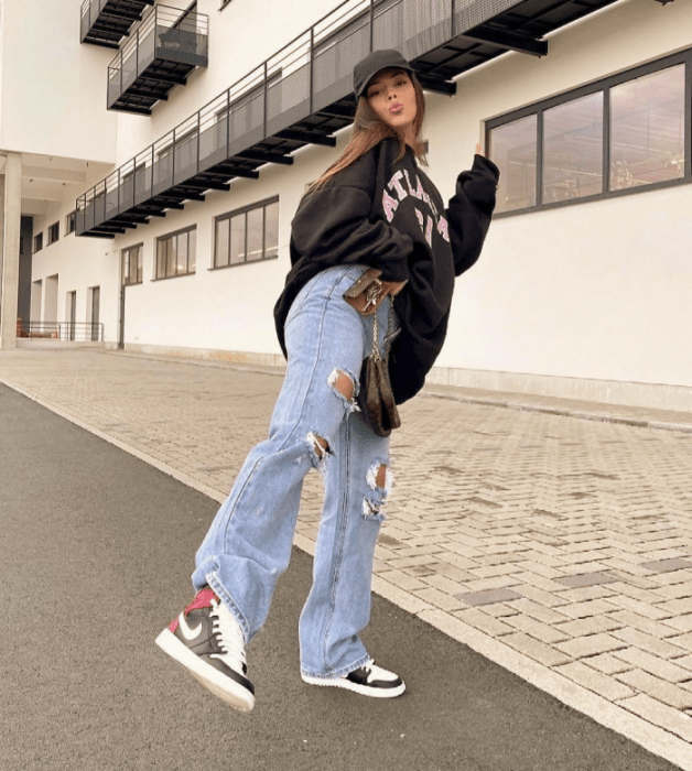 chica de cabello castaño con sudadera negra oversized, jeans rotos holgados, tenis blancos con negro y bucket hat negro