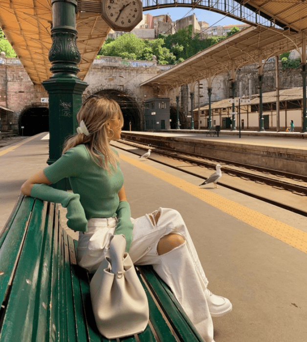 chica rubia con top verde tejido de manga larga, jeans blancos rotos, tenis blancos, bolso blanco grande de cuero
