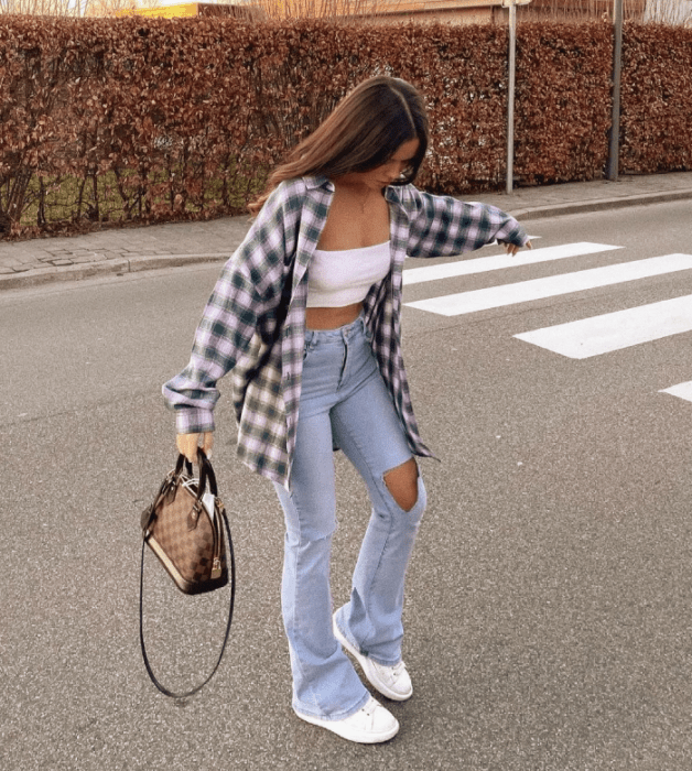chica de cabello castaño usando un crop top blanco, camisa de cuadros holgada, jeans acampanados rotos, tenis blancos y bolso café de piel