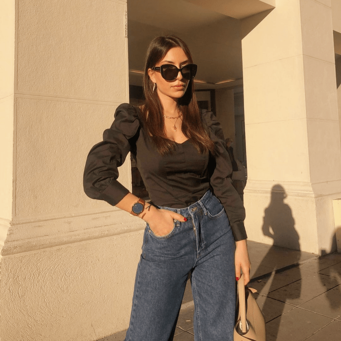 chica de cabello castaño usando lentes de sol, blusa negra de satén de manga larga y jeans ajustados