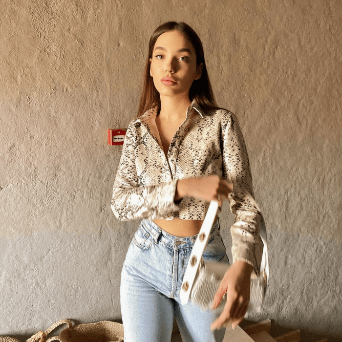 chica de cabello castaño usando una camisa crop top de satén con jeans claros y bolso blanco pequeño