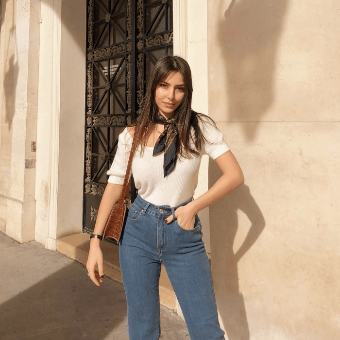 chica de cabello castaño usando una blusa blanca de manga 3/4, jeans ajustados a la cintura, bolsa café de cuero