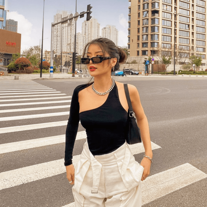 chica de cabello oscuro usando lentes de sol, una blusa negra asimétrica, pantalones blancos de vestir a la cintura, bolso pequeño negro
