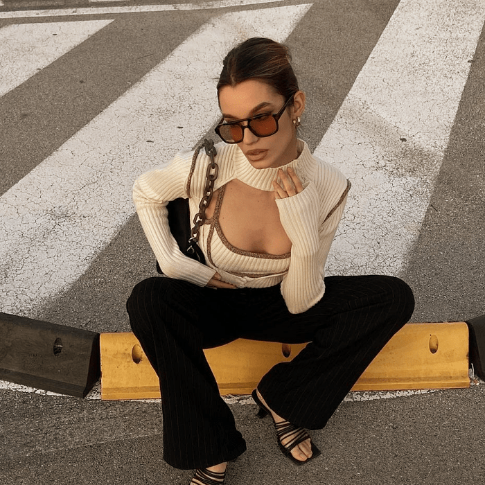 chica de cabello oscuro usando lentes de sol naranjas, blusa beige con orilla café, escote abierto, pantalones negros acampanados, sandalias negras de tacón