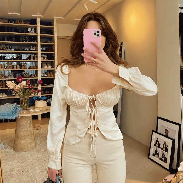 chica de cabello castaño usando una blusa beige de satén de manga larga con escote pronunciado y lazos al frente, jeans beige ajustados a la cintura