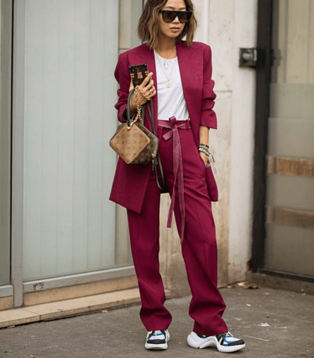 chica de cabello claro corto usando lentes de sol, camiseta blanca, traje fucsia, tenis blancos con negro y azul, bolso de mano café de piel
