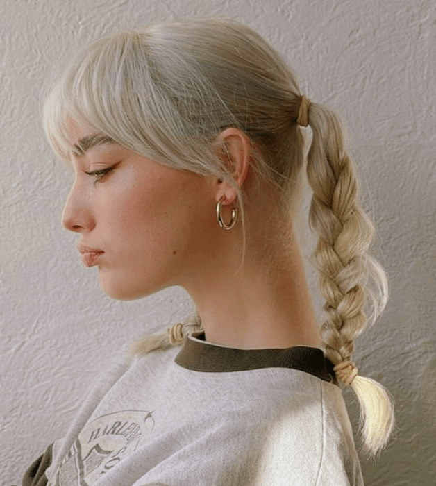 mujer de cabello platinado con coletas y trenzas, usando una camiseta blanca con cuello verde militar