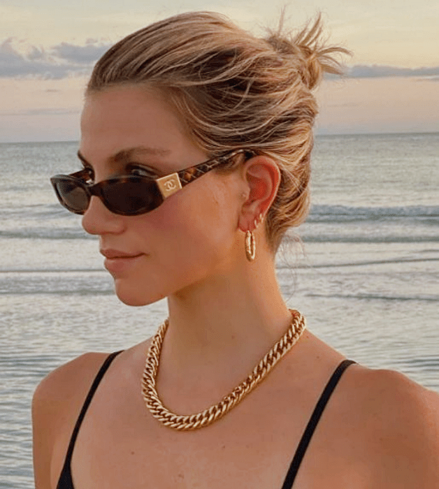 mujer rubia con gafas de sol, cabello recogido en un chongo detrás, collar dorado y blusa negra de tirantes