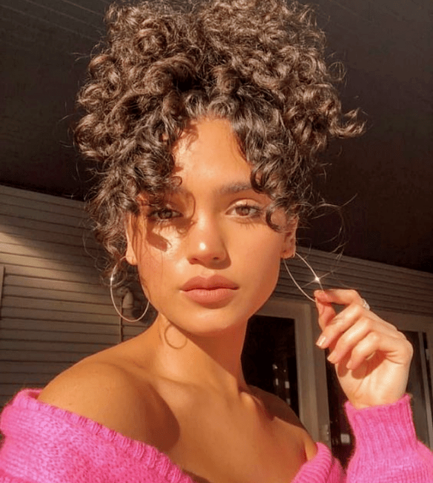 mujer de cabello moreno rizado usando un suéter rosa, con el cabello recogido en la parte alta