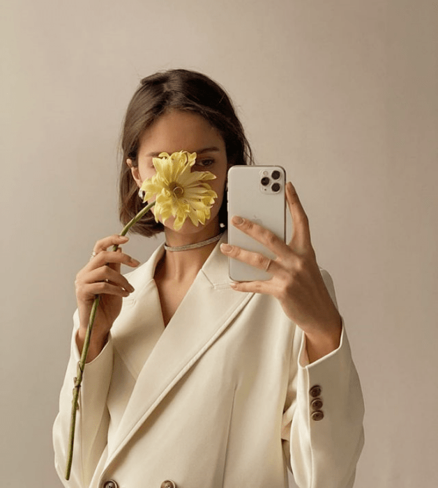 chica de cabello castaño corto usando un blazer grande beige con flor amarilla en la mano cubriendo su rostro