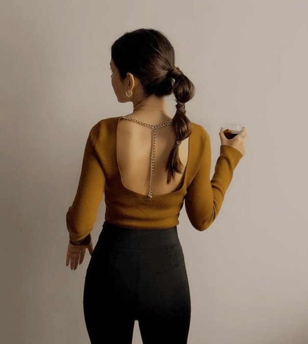 chica de cabello castaño con coleta, top mostaza de manga larga y espalda descubierta, jeans negros ajustados