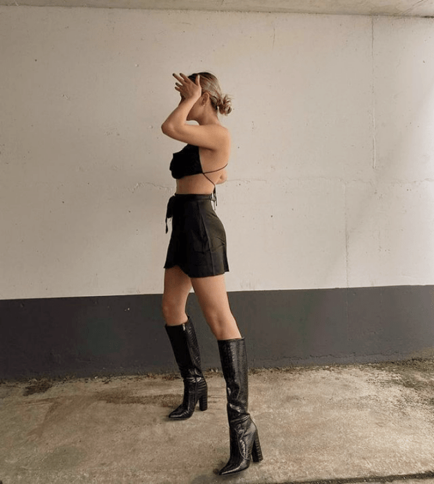 chica rubia usando minifalda negra, top negro de tirantes, botas largas de cuero