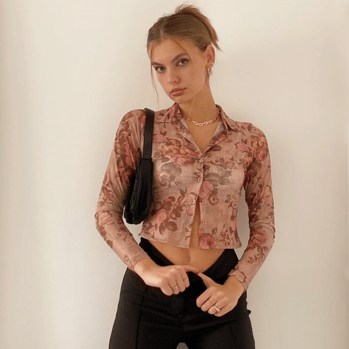 chica rubia usando un top semitransparente rosa con estampado floral, manga larga, jeans negros a la cintura y bolso negro de cuero