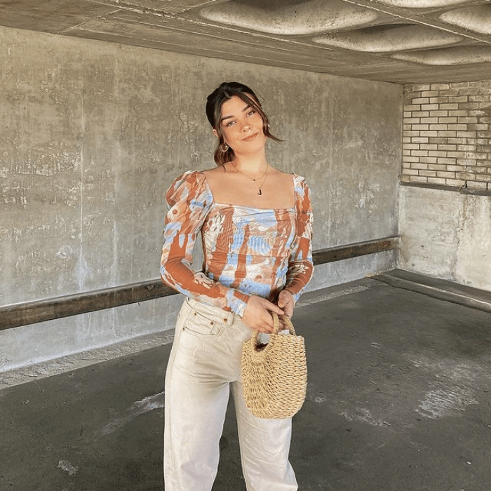 chica de cabello castaño usando un top celeste con líneas naranjas, pantalones beige a la cintura y bolso de mano pequeño beige