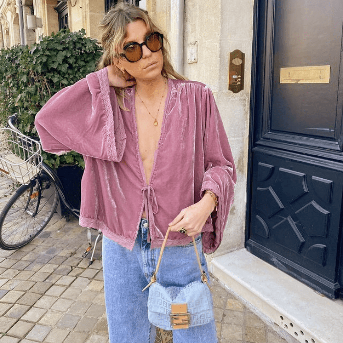 chica rubia con lentes de sol, blusa rosa de terciopelo con escote pronunciado, jeans a la cintura y bolso celeste con café