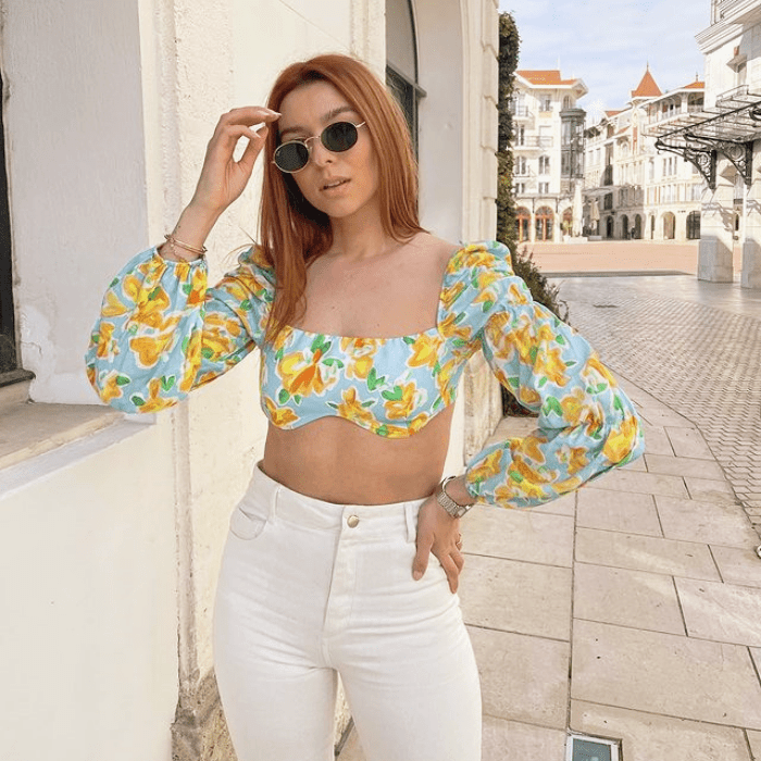 chica de cabello pelirrojo usando lentes de sol, blusa azul claro con flores amarillas, jeans a la cintura blancos