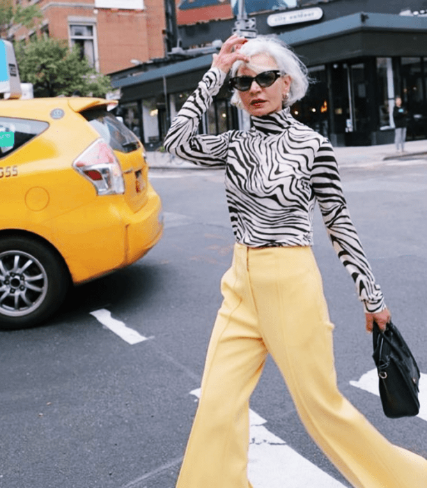 mujer mayor usando lentes de sol, cabello blanco, blusa de cebra de manga larga, pantalones amarillos acampanados y bolso negro de cuero