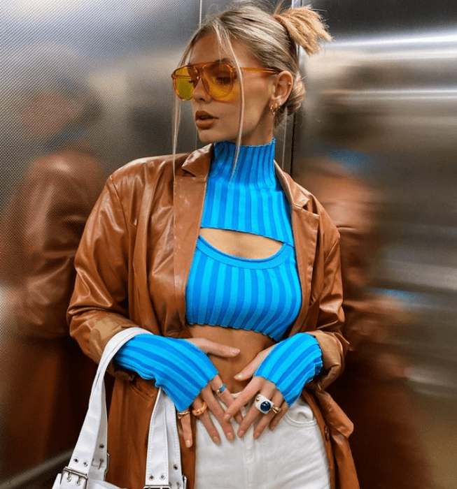 chica rubia con lentes de sol, top azul brillante con manga larga y escote asimétrico, pantalones blancos, abrigo de piel café y bolso blanco
