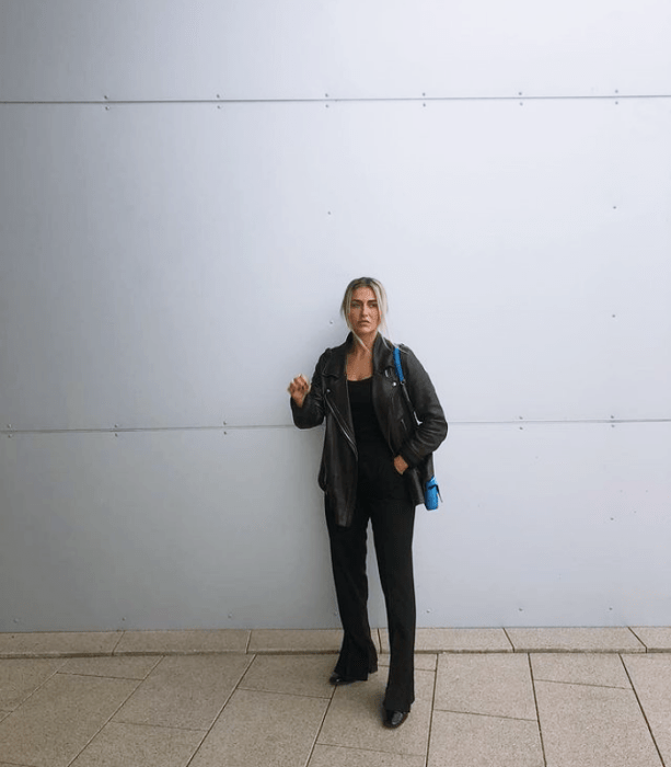 chica rubia usando una blusa negra, chamarra negra de cuero, jeans negros acampanados, botas negras, bolso negro