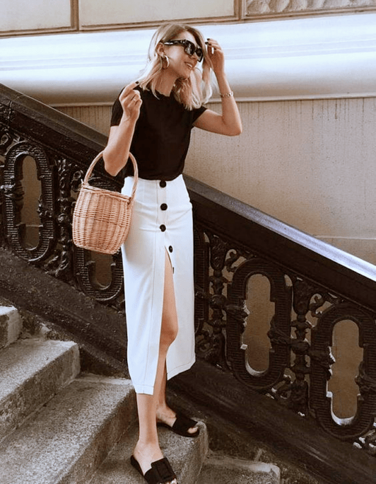 chica rubia usando un top negro de manga corta, falda larga blanca abierta con botones negros, flats negros