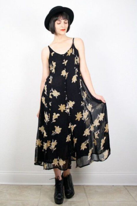 Chica usando un vestido largo de tirantes negros con estampado de flores amarillas, botines negros y sombrero negro