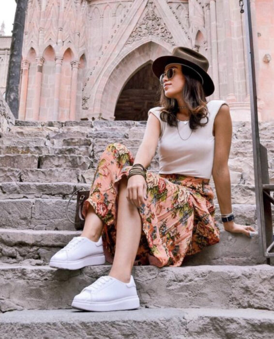 Chica usando blusa blanca con falda naranja y detalles verdes, tenis blancos y sombrero negro