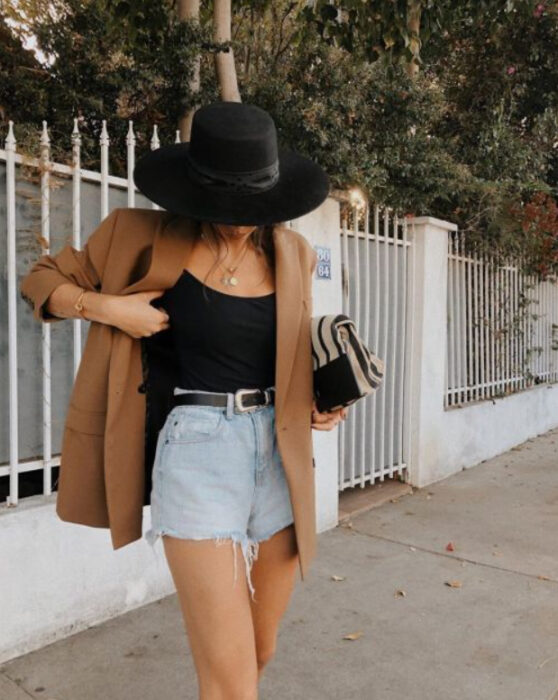 Chica usando blusa de tirantes negra, short de mezclilla clara, cinturón, blazer camel y sombrero negro